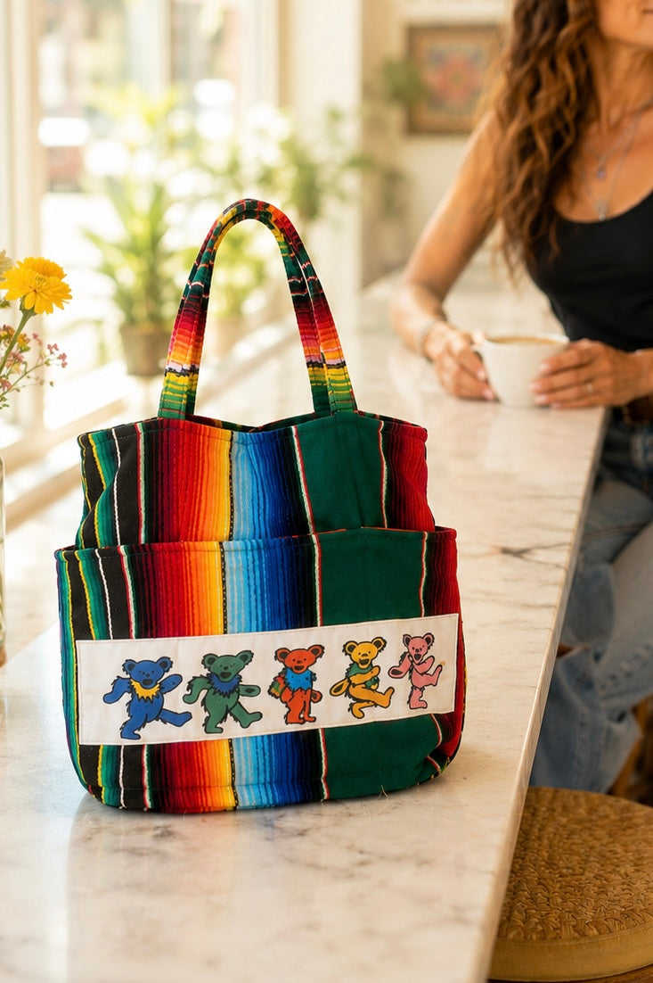A colorful striped tote bag with a white band featuring the Grateful Dead-style teddy bear logo, set on a marble counter in a bright cafe.