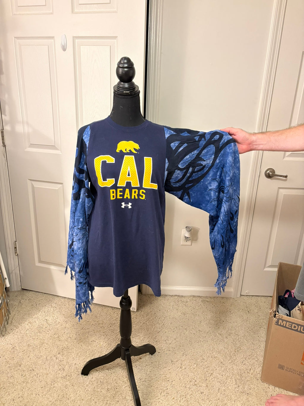 A navy blue long-sleeve t-shirt with 'CAL BEARS' and a bear logo printed on the front, displayed on a black dress form. The sleeves are attached to blue and black tie-dye fringed poncho-style overlays.
